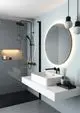 Waschtisch mit weißer quadratischer Waschschüssel auf minimalistischer Ablage, dazu eine schwarze Wand-Waschtischarmatur von Ideal Standard und runde beleuchtete Wandspiegel.